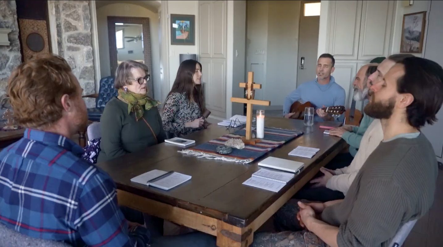 Guests and staff gathered around the table for worship, with a cross, candle, and guitar