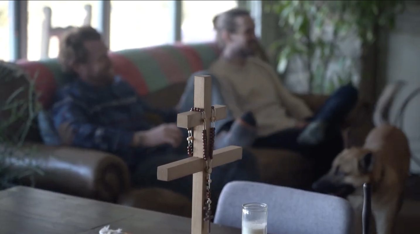 A handmade wooden cross with prayer beads on the table while guests rest in the living room